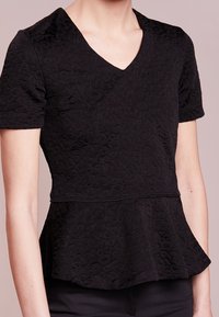 Musta peplum-toppi, joka on valmistettu tekstuuripintaisesta kankaasta, V-pääntiellä ja lyhyillä hihoilla. Ominaisuuksiin kuuluu istuva yläosa ja levenevä helma.