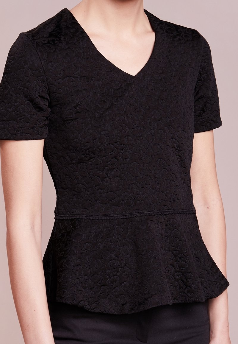 Musta peplum-toppi, joka on valmistettu tekstuuripintaisesta kankaasta, V-pääntiellä ja lyhyillä hihoilla. Ominaisuuksiin kuuluu istuva yläosa ja levenevä helma.