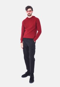 Maglione rosso strutturato con cappuccio, abbinato a pantaloni cargo neri e scarpe nere. Gli accessori includono occhiali da sole rosa. Outfit semplice e casual.