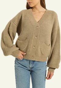 Eine Frau trägt eine beigefarbene, grob gerippte Strickjacke mit großen Knöpfen und Fronttaschen, kombiniert mit hellblauen Jeans.