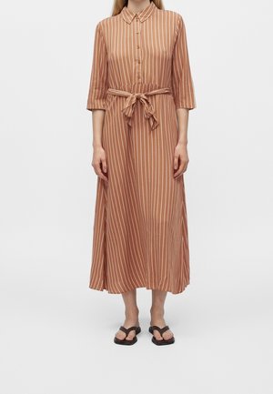 Femme portant une robe midi à rayures verticales marron et blanches, manches 3/4, devant boutonné, taille nouée, et sandales plates noires.