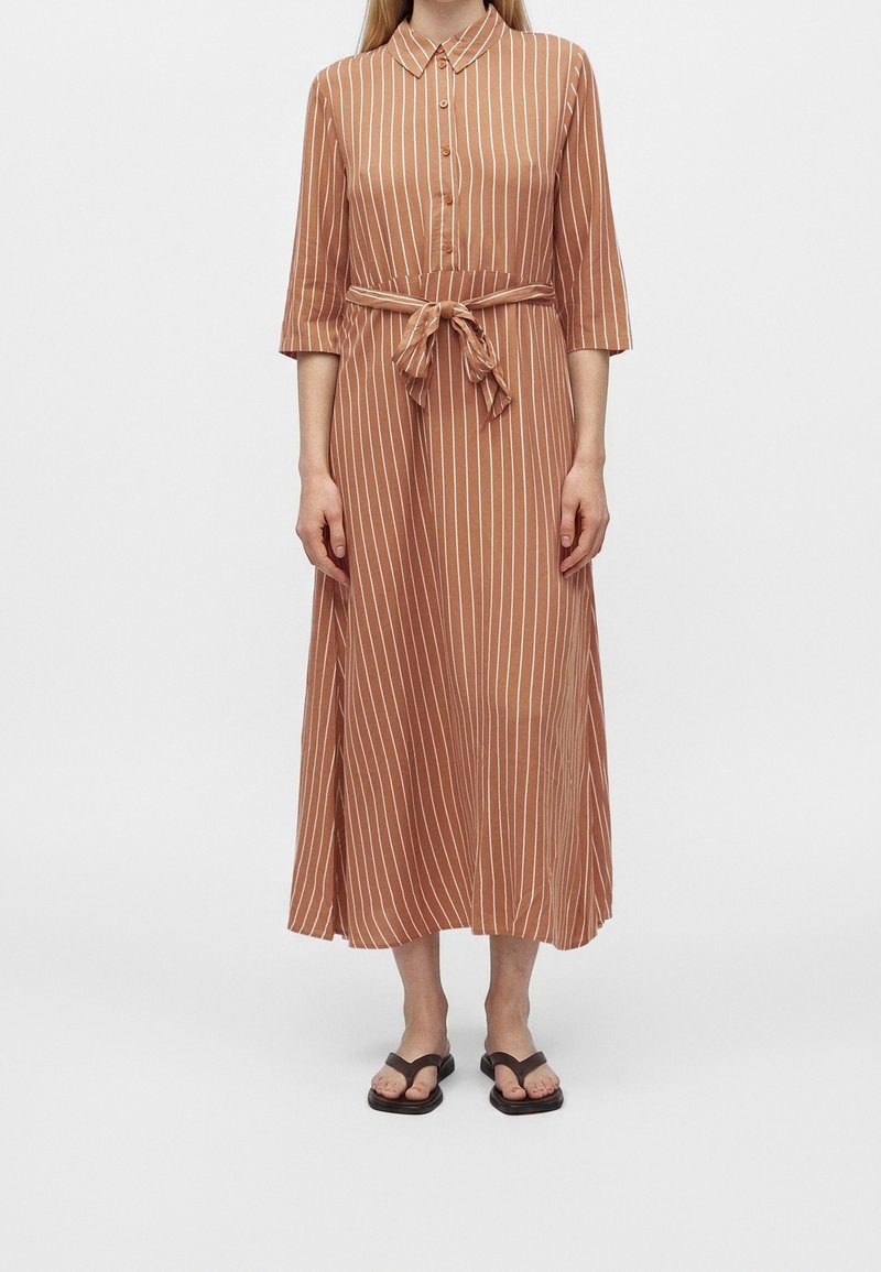 Femme portant une robe midi à rayures verticales marron et blanches, manches 3/4, devant boutonné, taille nouée, et sandales plates noires.