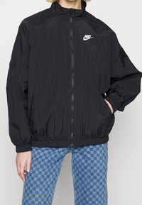 Person som bär en svart Nike zip-up jacka med elastiska ärmar och nederkant, i kombination med blå rutiga jeans.