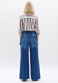 Jeans de mezclilla anchos de color azul con una textura desvanecida y dos bolsillos traseros. Combinados con una camisa a rayas con líneas verticales de múltiples colores.