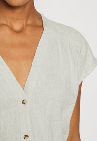 Ljusgrön linnéblus med V-ringning, korta ärmar och träknappar framtill. En svag struktur är synlig.