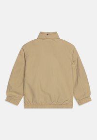 Beige lättviktsjacka med elastiska ärmslut och nederkant, avslappnad krage, broderad varumärkesnamn på ryggen. Slät textur, minimal design.