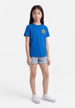 Jong meisje staand, gekleed in een blauw Santa Cruz T-shirt, lichtblauwe spijkershort, witte sokken en witte sneakers tegen een effen achtergrond.