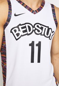 Maillot de basket-ball blanc avec le texte "BED-STUY", numéro 11, bordure à motifs multicolores, et logo Nike noir sur le haut à gauche de la poitrine.