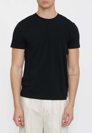 Uomo che indossa una maglietta nera semplice a maniche corte e pantaloni beige larghi, in piedi davanti a uno sfondo bianco semplice.