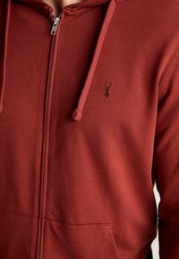 Sudadera con cremallera roja fabricada en una tela ligera, que cuenta con un bolsillo canguro, capucha con cordón y un pequeño logotipo bordado en el pecho.
