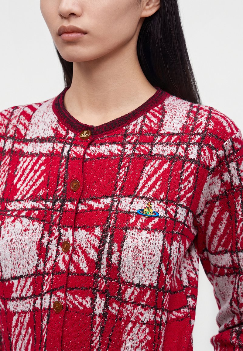 Femme portant un cardigan à carreaux rouge et blanc boutonné avec un petit logo coloré brodé sur la poitrine gauche.