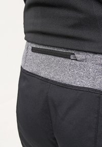 Primer plano de una persona con pantalones deportivos negros y grises que tienen un bolsillo con cremallera cerca de la cintura y una camiseta negra texturizada.