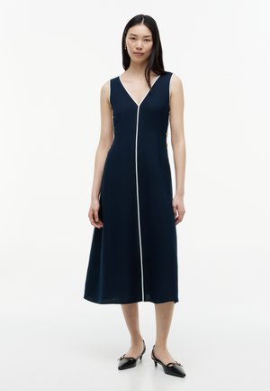 Mujer que lleva un vestido midi azul marino sin mangas con ribete blanco por el centro y botones dorados en los laterales, combinado con zapatos de tacón bajos negros.