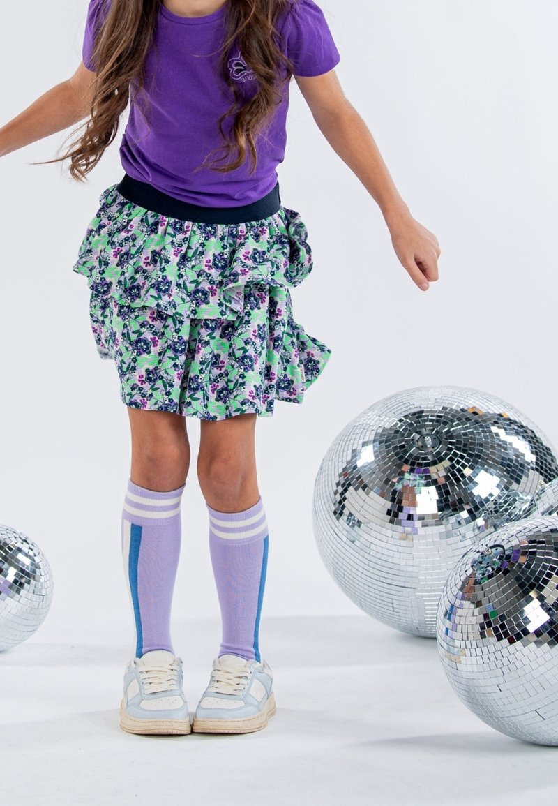 Enfant portant une chemise violette, une jupe fleurie et des chaussettes montantes se tient parmi trois grandes boules disco réfléchissantes sur un sol blanc.