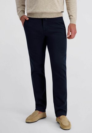 Man die marineblauwe broek, beige loafers en een beige trui draagt, met één hand in de zak, staande tegen een effen achtergrond.