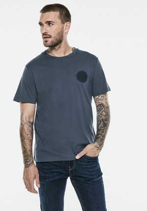 T-shirt in cotone grigio scuro con scollo rotondo, maniche corte e una piccola grafica nera sul petto, abbinata a jeans blu.