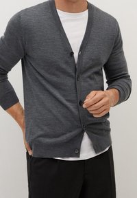 Mann trägt eine graue, geknöpfte Strickjacke über einem weißen Hemd und schwarzen Hosen, richtet die Strickjacke mit einer Hand.