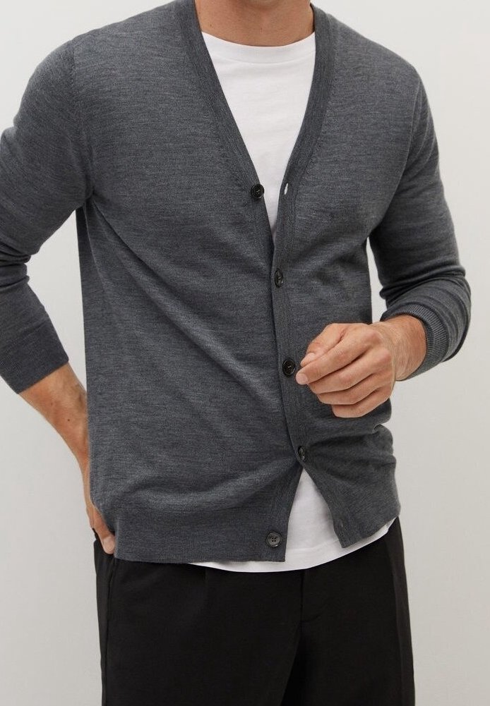 Mann trägt eine graue, geknöpfte Strickjacke über einem weißen Hemd und schwarzen Hosen, richtet die Strickjacke mit einer Hand.