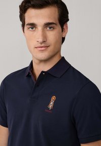 Marinblå polo skjorta i bomull med strukturerad yta, med en broderad design och logotyp på vänster bröst.
