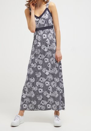 Kvinde iført en ærmeløs sort og hvid blomstret maxikjole med V-udskæring og hvide sneakers, stående foran en ensfarvet baggrund.