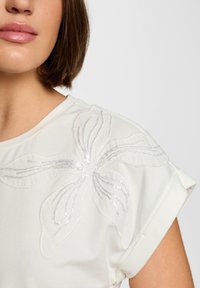 White short-sleeve blouse featuring a floral design in silver sequins on the shoulder, with a smooth texture and rolled cuff detail.