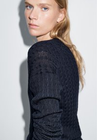 Junge Frau mit heller Haut und blondem Haar trägt einen dunkelblauen Strickpullover mit Zopfmuster und schaut über ihre Schulter vor einem einfarbigen Hintergrund.