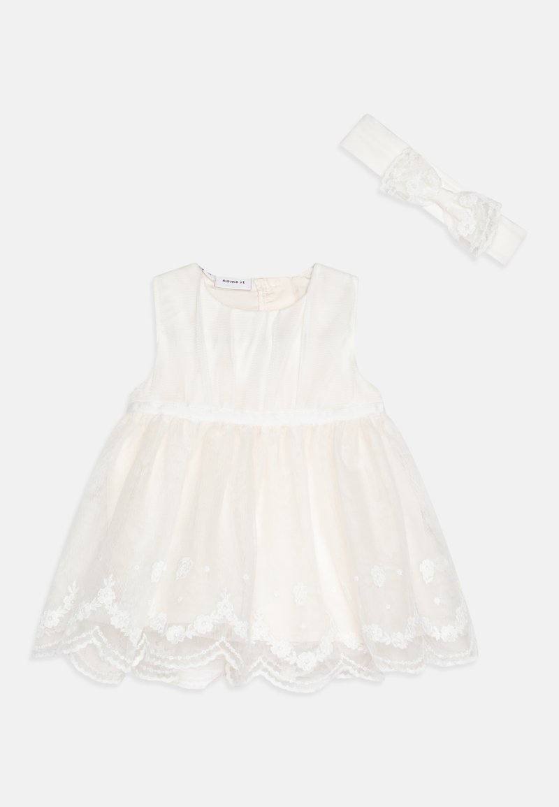 Balta siuvinėta suknelė su plačiu sijonu ir nėrinių apdaila, kartu su atitinkama galvos juosta, kurioje yra kaspinėlis ir nėrinių akcentai.