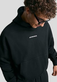 Čierna mikina s kapucňou, mäkká tkanina, voľný strih, obsahuje predné vrecko a biele logo "OVRNRM" na hrudi.