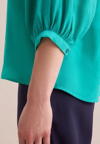 Blusa teal con maniche a sbuffo e dettagli conbottoni ai polsi, realizzata in tessuto leggero; abbinata a pantaloni blu scuro.