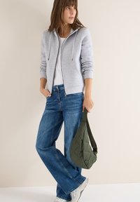 Světlá šedá zipová mikina s tkaničkami, bílý tričko, volné modré džíny a prošívaná zeleno-olivová přes rameno. Stříbrné tenisky.