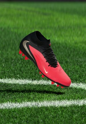Κόκκινο και μαύρο ποδοσφαιρικό παπούτσι Nike με χρυσό λογότυπο, αιωρούμενο πάνω από πράσινο γρασίδι με λευκές γραμμές γηπέδου.