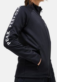 Giacca nera con zip e colletto alto, realizzata in tessuto liscio. Presenta il logo "PEAK PERFORMANCE" lungo la manica destra e tasche laterali.