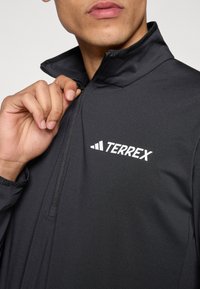 Melnā Terrex jaka ar augstu apkakli, puszipu dizainu, gludu tekstūru un baltu logo uz kreisās krūtis. Redzamas regulējamas aproces.