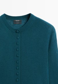 Turquoise, knoopvest gemaakt van 100% wol. Heeft een ronde halslijn en gelijkmatig verdeelde knopen bekleed met stof aan de voorkant.