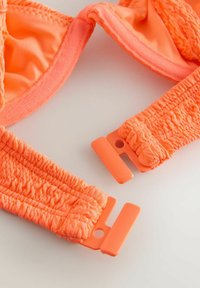 Heledad oranžid elastsed kangarihmad plastsest külgmise vabastusega pandlatega, mis asuvad heledal pinnal.