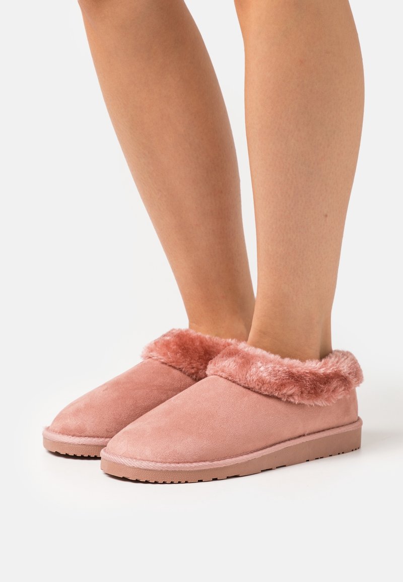 Pieds portant des chaussons roses duveteux à enfiler avec des semelles épaisses sur fond blanc.