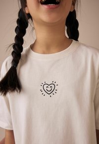 T-shirt en coton crème avec un dessin de cœur noir souriant entouré de rayons, manches courtes et col rond.