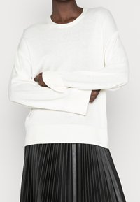 Vit långärmad tröja med rund halsringning och ribbade ärmslut. Över en svart plisserad kjol verkar tyget mjukt och strukturerat.