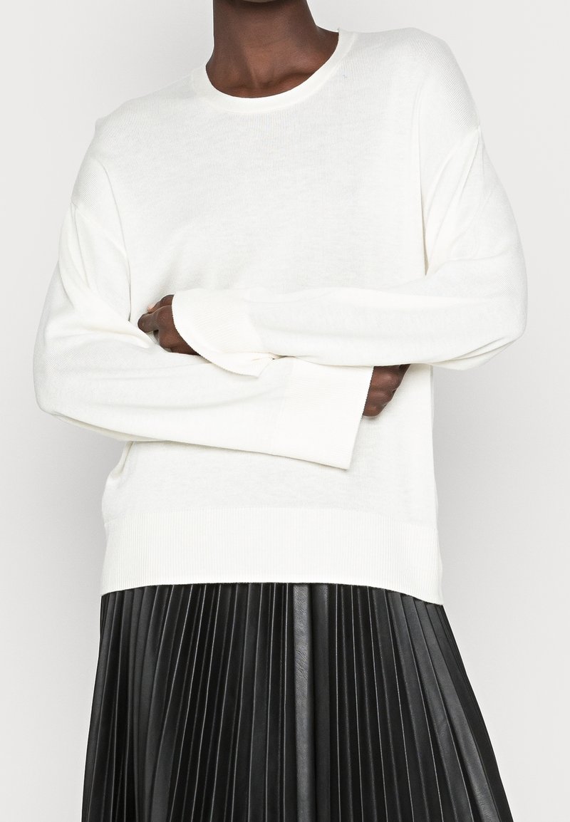 Vit långärmad tröja med rund halsringning och ribbade ärmslut. Över en svart plisserad kjol verkar tyget mjukt och strukturerat.