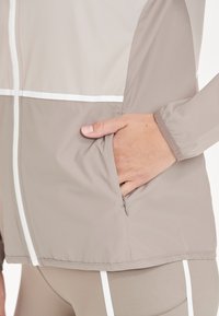 Lätt beige jacka med en zippered ficka, vita accentremsor och ett mjukt, texturerat tyg med en åtsittande ärmdesign.