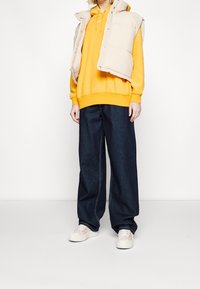 Beige toppaliivi keltaisen hupparin päällä, yhdistetty tummansinisiin leveälahkeisiin farkkuihin ja valkoisiin lenkkareihin, joissa on värikkäitä yksityiskohtia.