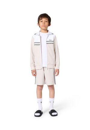 Garçon portant une veste beige zippée, un short assorti avec des bandes latérales, des chaussettes blanches, des sandales noires slides et un t-shirt blanc, debout sur un fond blanc.