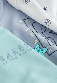 Tre T-shirts i pastelfarver: mintgrøn med logo, lyseblå med guldskrift og hvid med marineblåt blomsterprint.