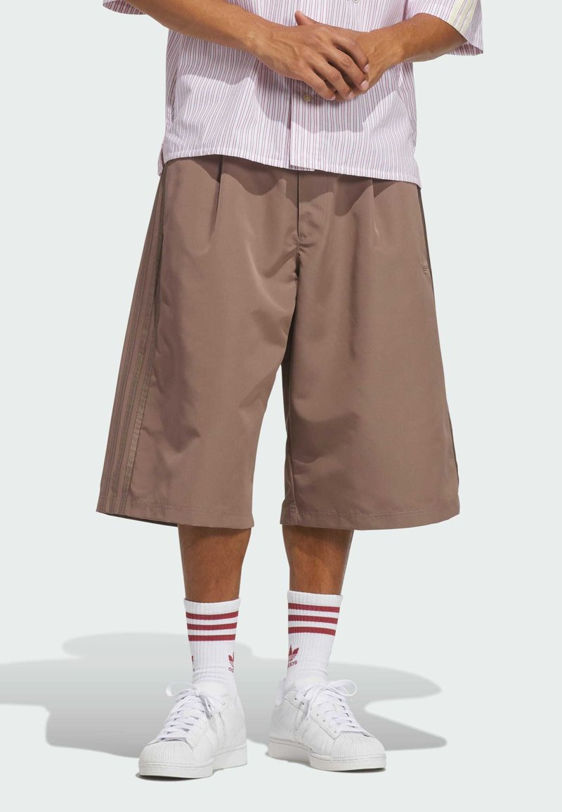 Person wearing brown loose knee-length shorts, white sneakers, and white socks with red stripes, standing with hands clasped.