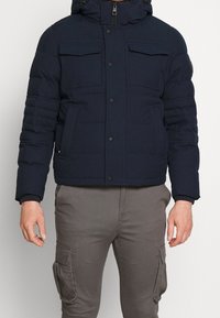 Granatowa kurtka puchowa z kapturem, dwoma kieszeniami na piersi oraz elastycznymi mankietami. Zapięcie na suwak i pikowany wzór. Luźny krój, na co dzień.