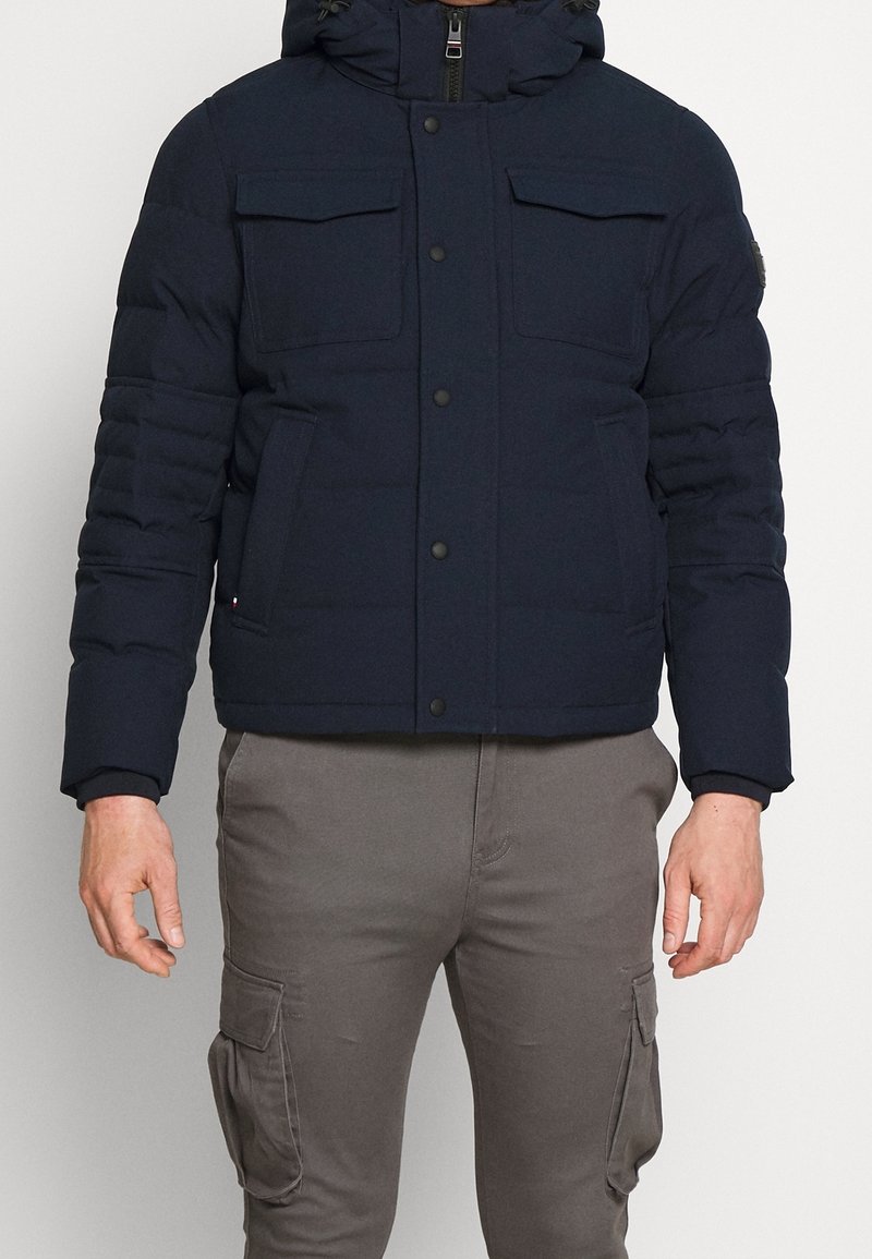 Granatowa kurtka puchowa z kapturem, dwoma kieszeniami na piersi oraz elastycznymi mankietami. Zapięcie na suwak i pikowany wzór. Luźny krój, na co dzień.