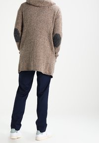 Persona che indossa un maglione a maglia beige con toppe grigie sui gomiti, pantaloni navy e sneakers bianche in piedi contro uno sfondo semplice.