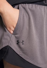 Mano descansando dentro del bolsillo lateral de unos pantalones cortos deportivos Under Armour grises con ribete negro.