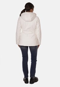 Giacca impermeabile bianca con cappuccio, caratterizzata da un esterno liscio, design aderente e maniche lunghe. Indossata sopra jeans scuri e stivali.