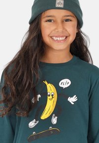 Långärmad tröja i teal med en banan grafik som har armar och ben och åker skateboard, åtföljd av ordet "FLIP" i en pratbubbla.